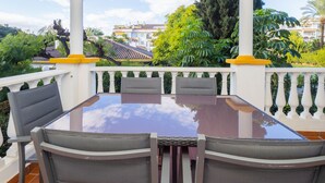 Un espacio cubierto para comer al aire libre con una mesa de vidrio y sillas de mimbre, rodeado por un balcón blanco y detalles en amarillo, con vistas a un barrio residencial.