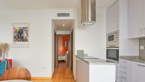 Eine moderne Küche mit Einbauofen und Mikrowelle, ein Essbereich mit Holztisch und Stühlen sowie ein Flur, der zu einem Schlafzimmer mit orangefarbener Akzentwand führt.
