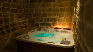 Ein Zimmer mit Steinwänden, einer Holztonne gefüllt mit Wasser und Blasen, sowie einem kleinen Tisch mit Kerzen und einem Getränk.