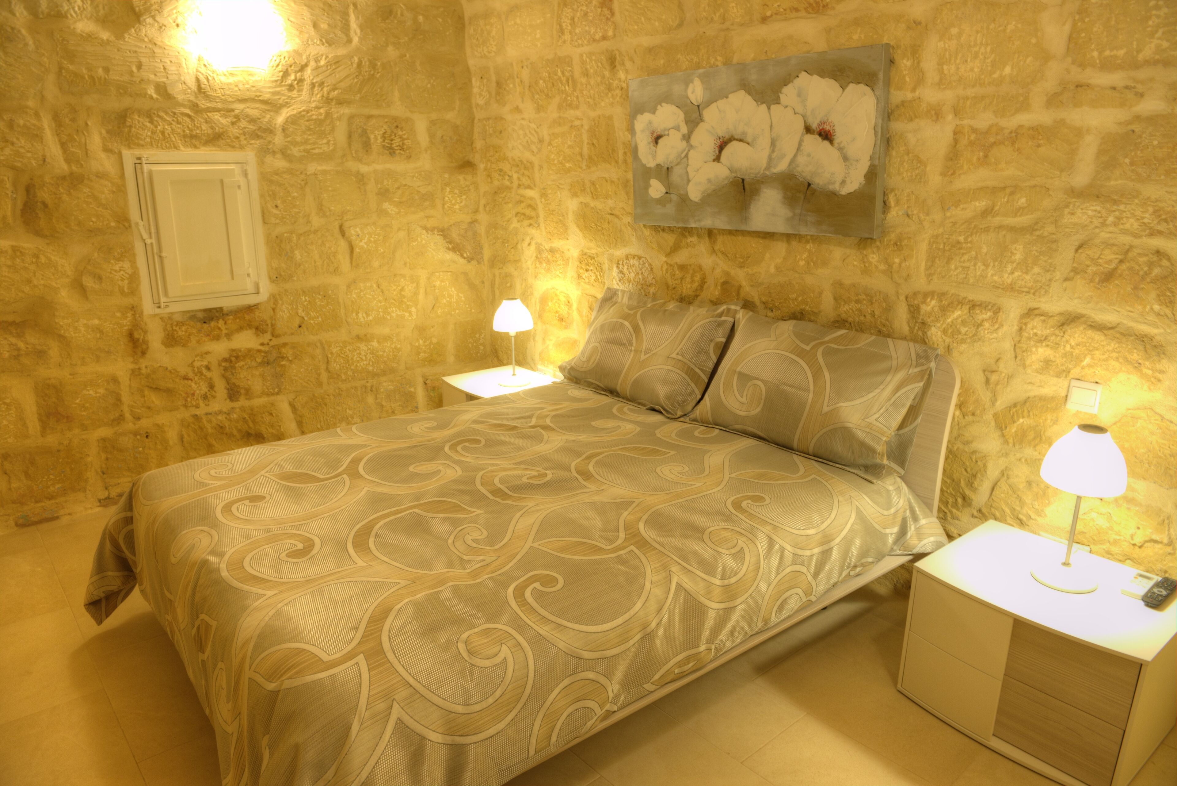 A bedroom with a stone wall, a bed with a patterned bedspread, two bedside tables with lamps, and a floral painting on the wall.