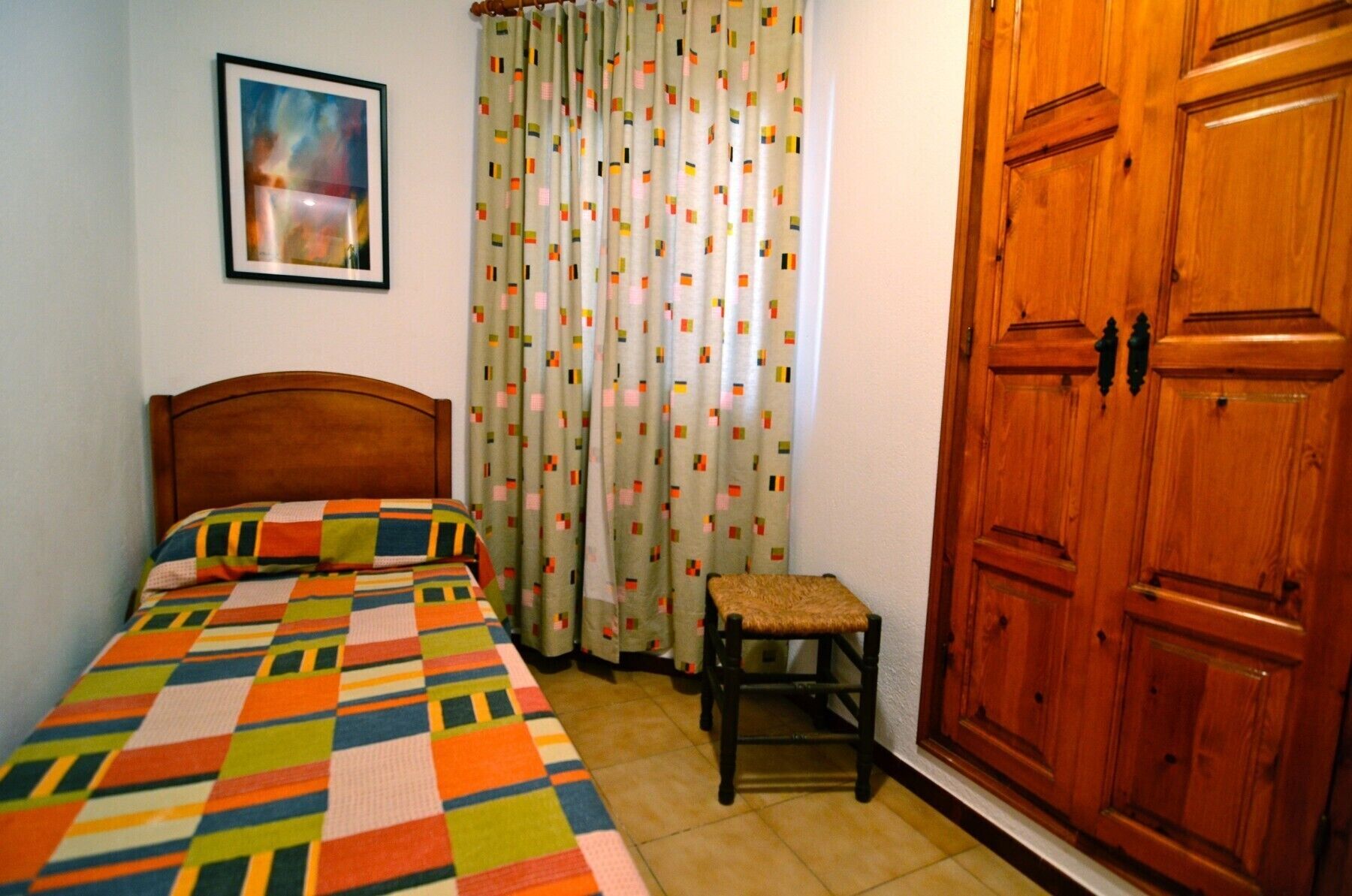 Un dormitorio con una cama de madera, un edredón colorido, una silla, un armario de madera y una cortina estampada.