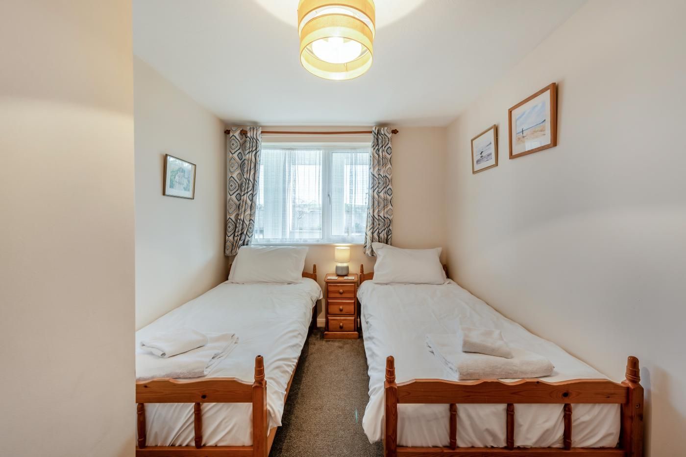 Een kamer met twee houten bedden, een raam met gordijnen met een patroon, een nachtkastje met een lamp, en lijstwerken aan de muur.