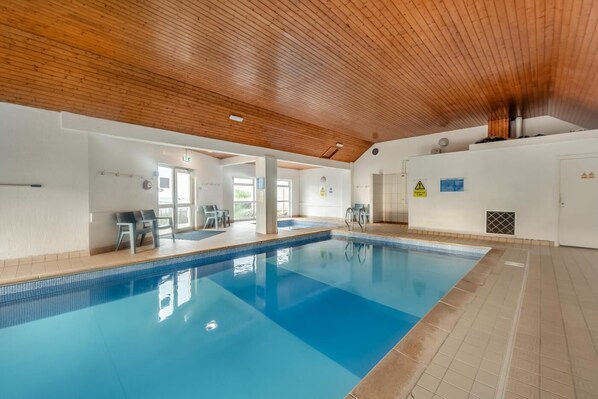 An indoor swimming pool with a wooden ceiling, clear glass walls, and a tiled floor.