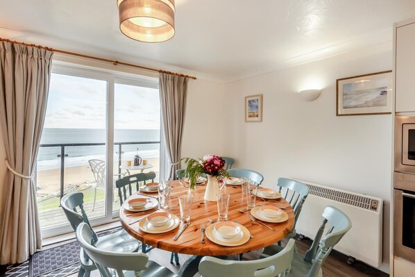 Comedor con una mesa redonda de madera dispuesta para cuatro personas, rodeada de sillas azules. Se ve una playa a través de la puerta de vidrio, y un radiador es visible en la pared.