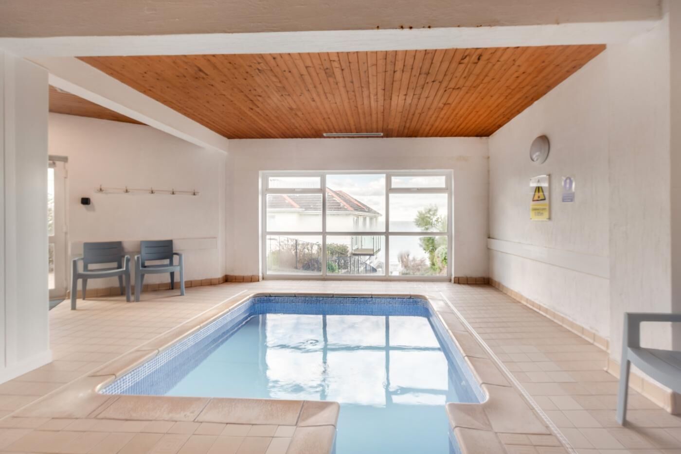 Una piscina amb sostre de fusta i terra de rajola, dues cadires i una finestra gran amb vista a una casa.