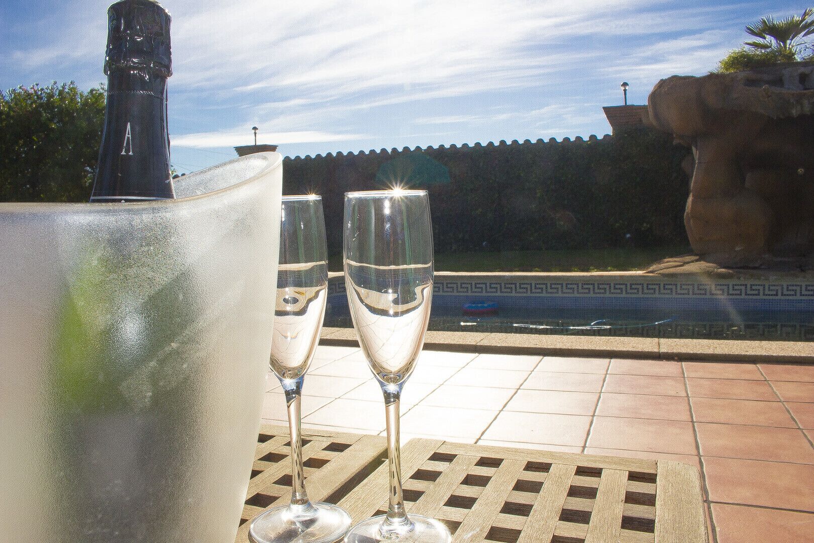 A poolside setting with two champagne flutes on a tiled table, a large ice bucket, and a stone structure in the background.