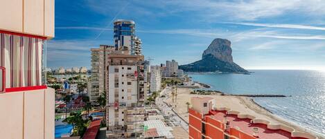 A coastal cityscape with a prominent mountain in the background, a beach, and various buildings including a tall structure with a blue top.