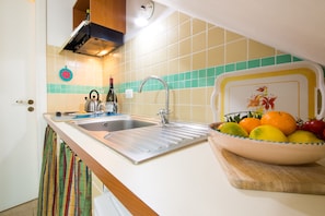 A kitchen with a stainless steel sink, a wooden cutting board with fruit, and a tiled backsplash. - Rooftop house in the center, 2/4 guests, free illimited WIFI, free parking (Scicli)