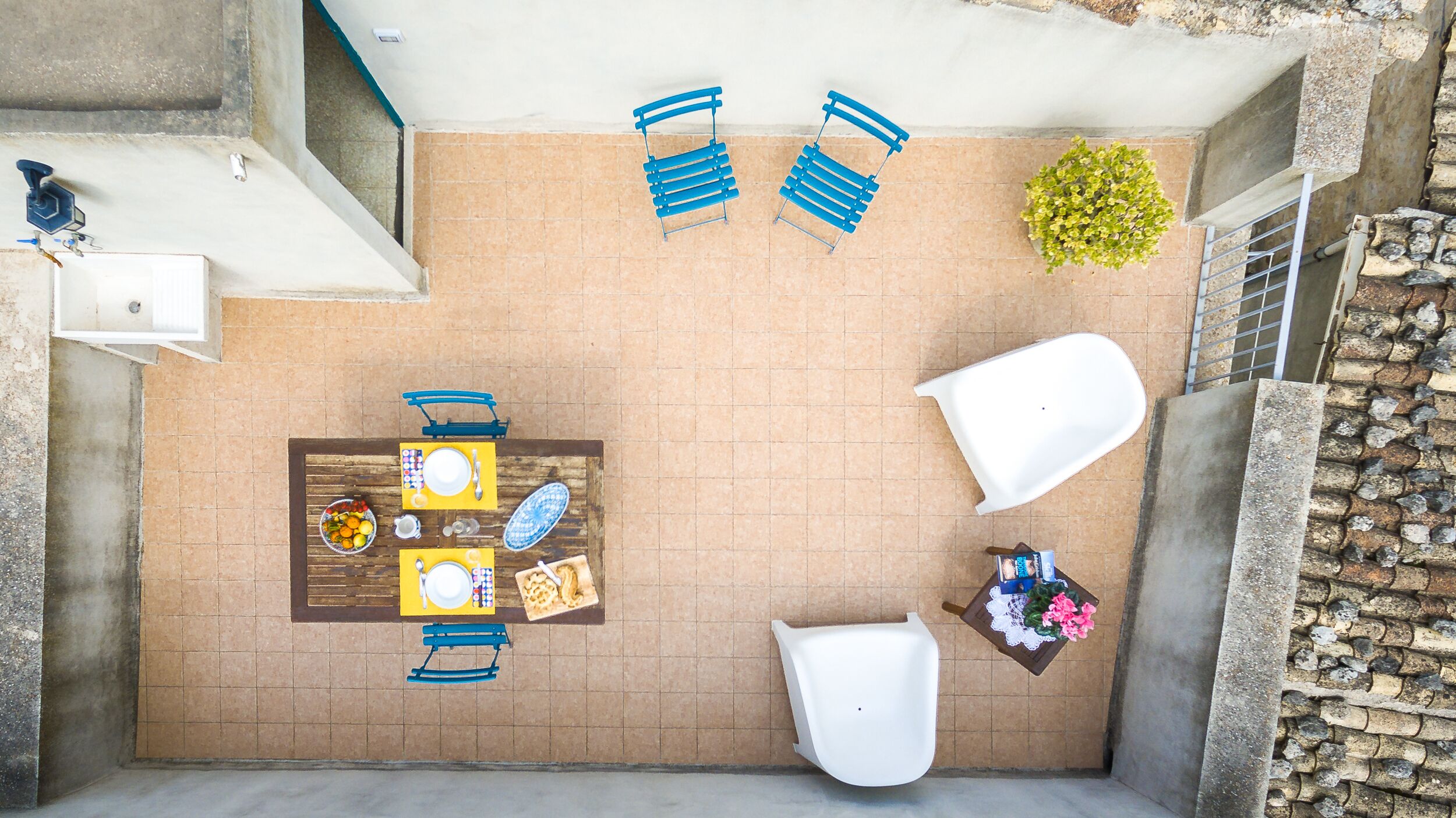 A patio with a table set for two, two blue chairs, a white chair, and a small white table. There is a potted plant and a white object on the right side.