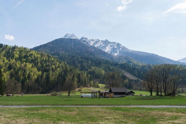 Paisaje rural con una casa y un campo en primer plano, una cadena montañosa al fondo y un cielo despejado.