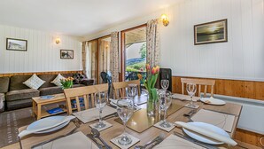 Un coin repas avec une table en bois dressée pour quatre personnes, entourée de chaises. Deux tableaux encadrés ornent les murs, il y a une fenêtre avec des rideaux et une vue sur l’extérieur.