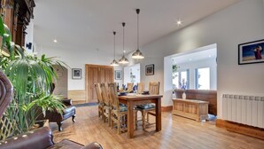 A spacious living room with a wooden dining table, chairs, and a sofa. There are pendant lights, a radiator, and a large window providing natural light. - 2 Bed in Lynton (oc-cslewt) (Lynmouth)