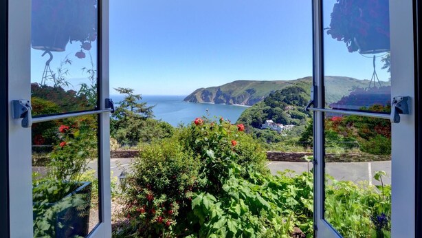 A view through a window with a garden in the foreground, a coastal landscape with hills and a body of water in the background. - 2 Bed in Lynton (oc-cslewt) (Lynmouth)