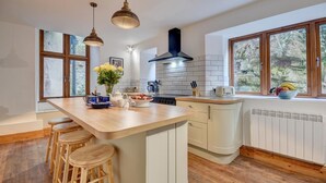 A modern kitchen with a large island, wooden stools, pendant lights, and a view of greenery through the windows. - 2 Bed in Lynton (oc-cslewt) (Lynmouth)