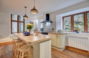 A modern kitchen with a large island, wooden stools, pendant lights, and a view of greenery through the windows.