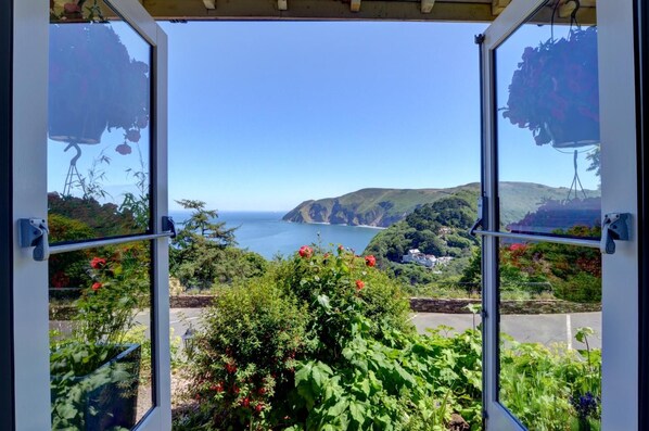 A view through a window with a garden in the foreground, a coastal landscape with hills and a body of water in the background.