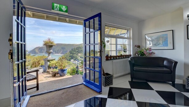 A room with a large open door leading to a balcony with a view of the sea and hills. There is a black and white checkered floor, a black sofa, and a radiator on the wall. - 2 Bed in Lynton (oc-cslewt) (Lynmouth)