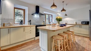 A modern kitchen with a central island, wooden stools, a black range hood, and pendant lighting. - 2 Bed in Lynton (oc-cslewt) (Lynmouth)