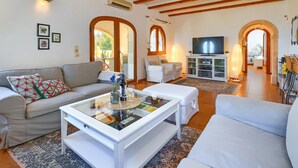 A spacious living room with a sofa, a glass coffee table, and a TV. There are two chairs and a cabinet with a TV. The room has wooden beams on the ceiling and a large window.