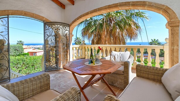 A balcony with wicker furniture, a table, and a view of the sea and palm trees.