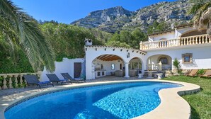 A swimming pool with lounge chairs, a patio area with outdoor furniture, and a mountainous backdrop.
