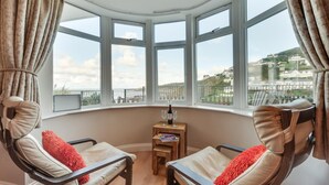 A corner room with large windows, two leather chairs, a small wooden table with a bottle and glasses, and a view of buildings and greenery. - 1 Bed in Woolacombe (oc-waves) (Woolacombe)