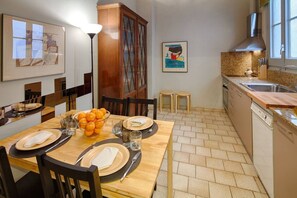 A dining area with a wooden table set for four, a fruit bowl, and a painting on the wall.