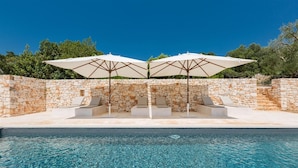 A swimming pool with clear blue water, surrounded by stone walls and two white umbrellas providing shade.