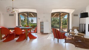 A spacious living area with a dining table and chairs, a striped armchair, and a large window offering a view of greenery.