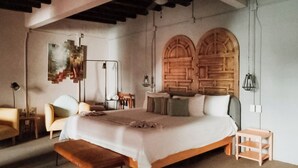Un dormitorio con una cama grande, dos puertas de madera en arco, un banco, una silla y una mesita.