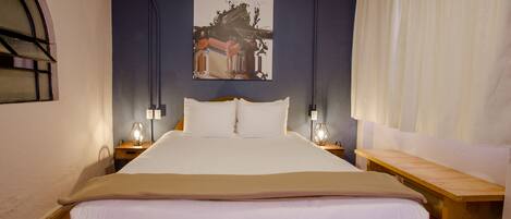 A neatly made bed with white linens, a wooden headboard, and a bench beside it. There is a wall-mounted artwork above the bed and a window with a curved design on the left.