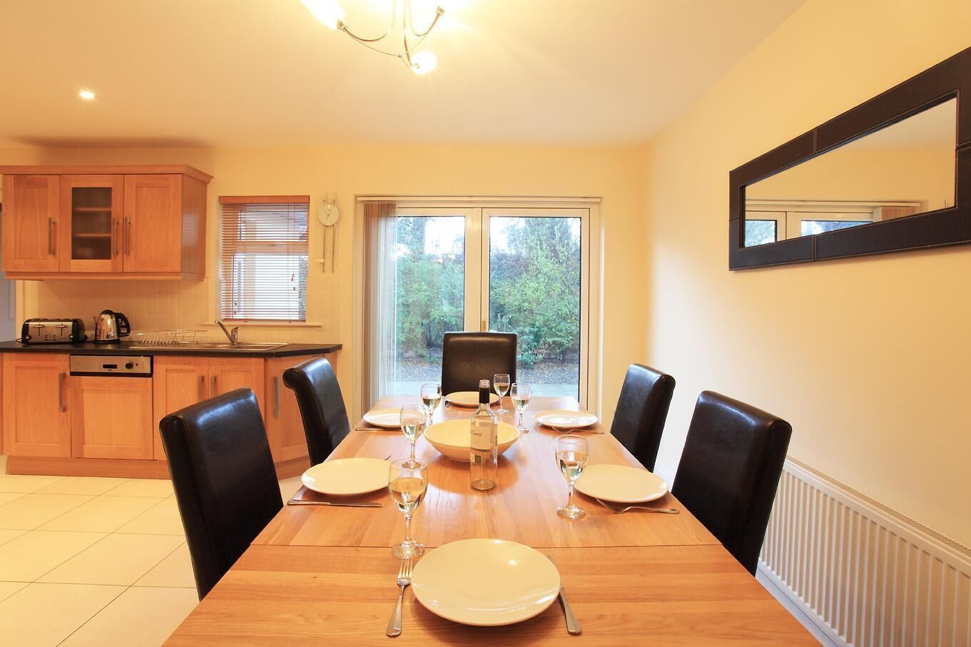 A dining area with a wooden table set for six, surrounded by black chairs. A kitchen with wooden cabinets and a window is visible in the background.