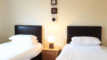 A hotel room with two beds, each with white bedding and a dark headboard. There is a nightstand between the beds with a lamp and a small clock on the wall above.