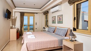 A hotel room with a large bed, a TV, an air conditioner, a window with curtains, and a view of the sea. - Palladio Luxury Villa with amazing view (Roussospiti)