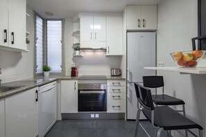 Une cuisine compacte avec des meubles blancs, un four encastré, un réfrigérateur et un petit coin repas avec deux chaises.