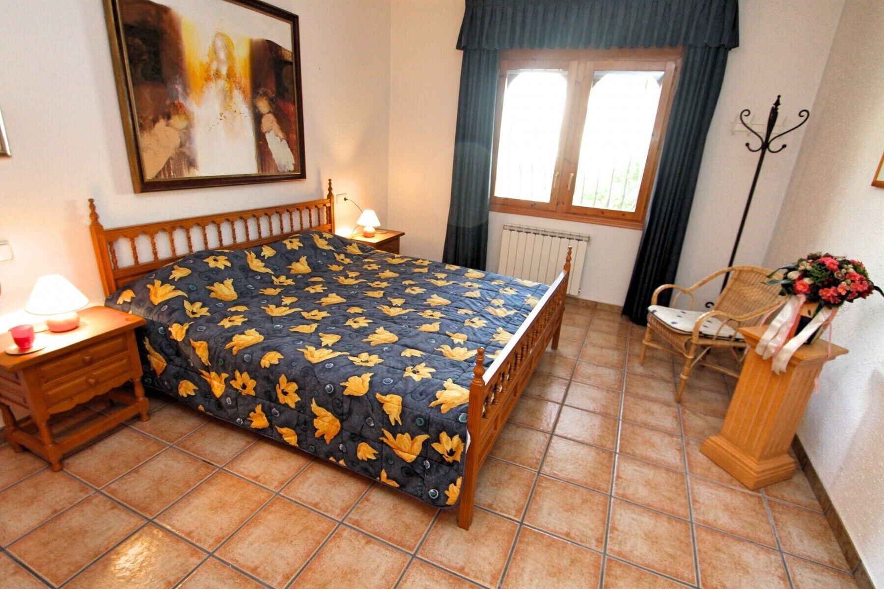 Un dormitorio con cama, mesita de noche, silla, ventana con cortinas y un cuadro en la pared.