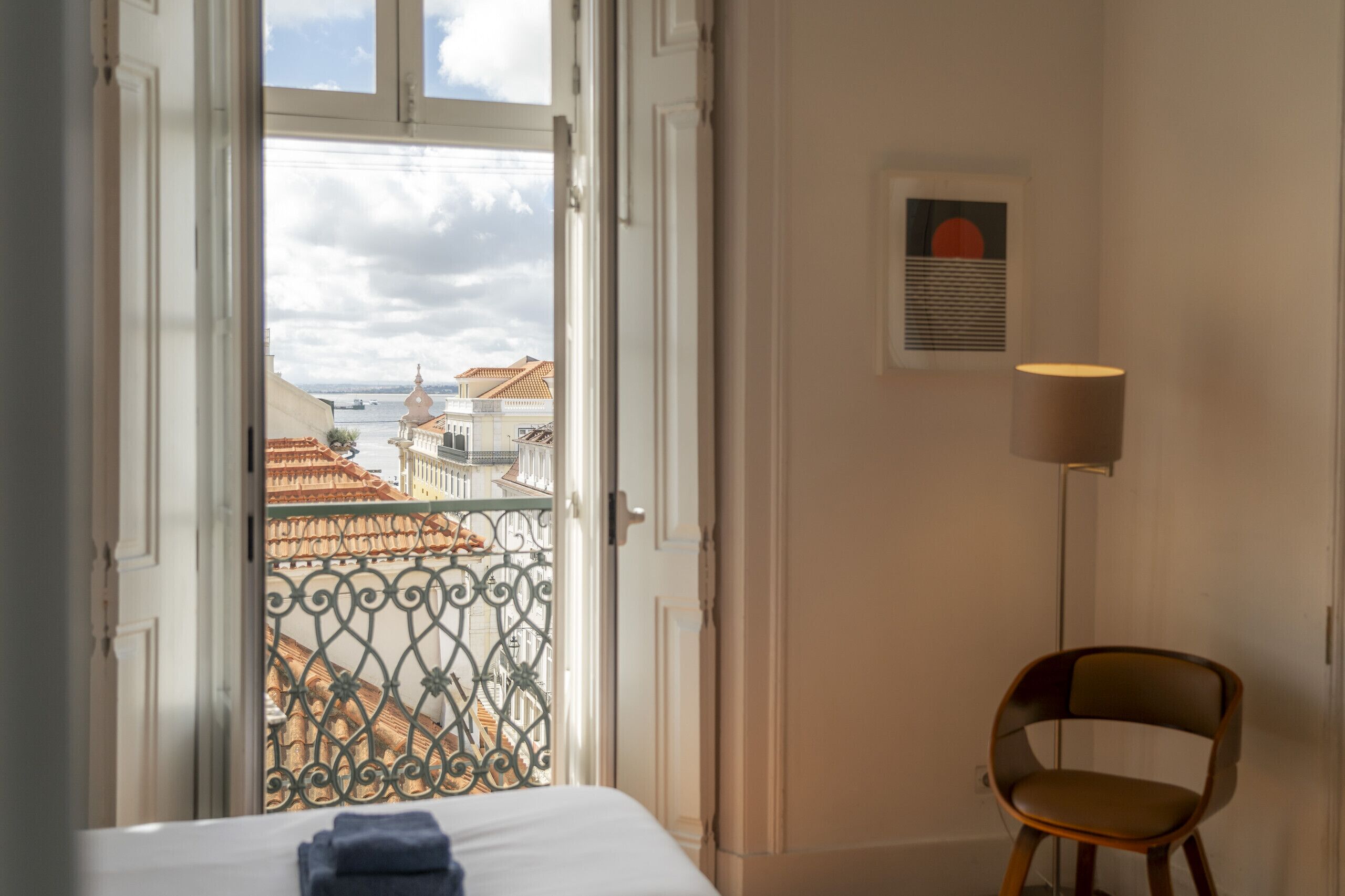 Une chambre d’hôtel avec un lit, une chaise, une lampe, une fenêtre donnant sur des immeubles et un tableau encadré au mur.
