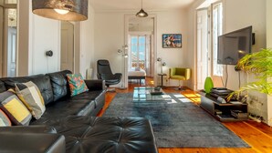 Un salon moderne avec un canapé en cuir noir, une table basse en verre, un grand luminaire suspendu noir, un téléviseur à écran plat et une plante en pot.