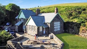 A stone building with a green roof, a stone patio, and a grassy lawn. - The School House, Countisbury (Lynton)