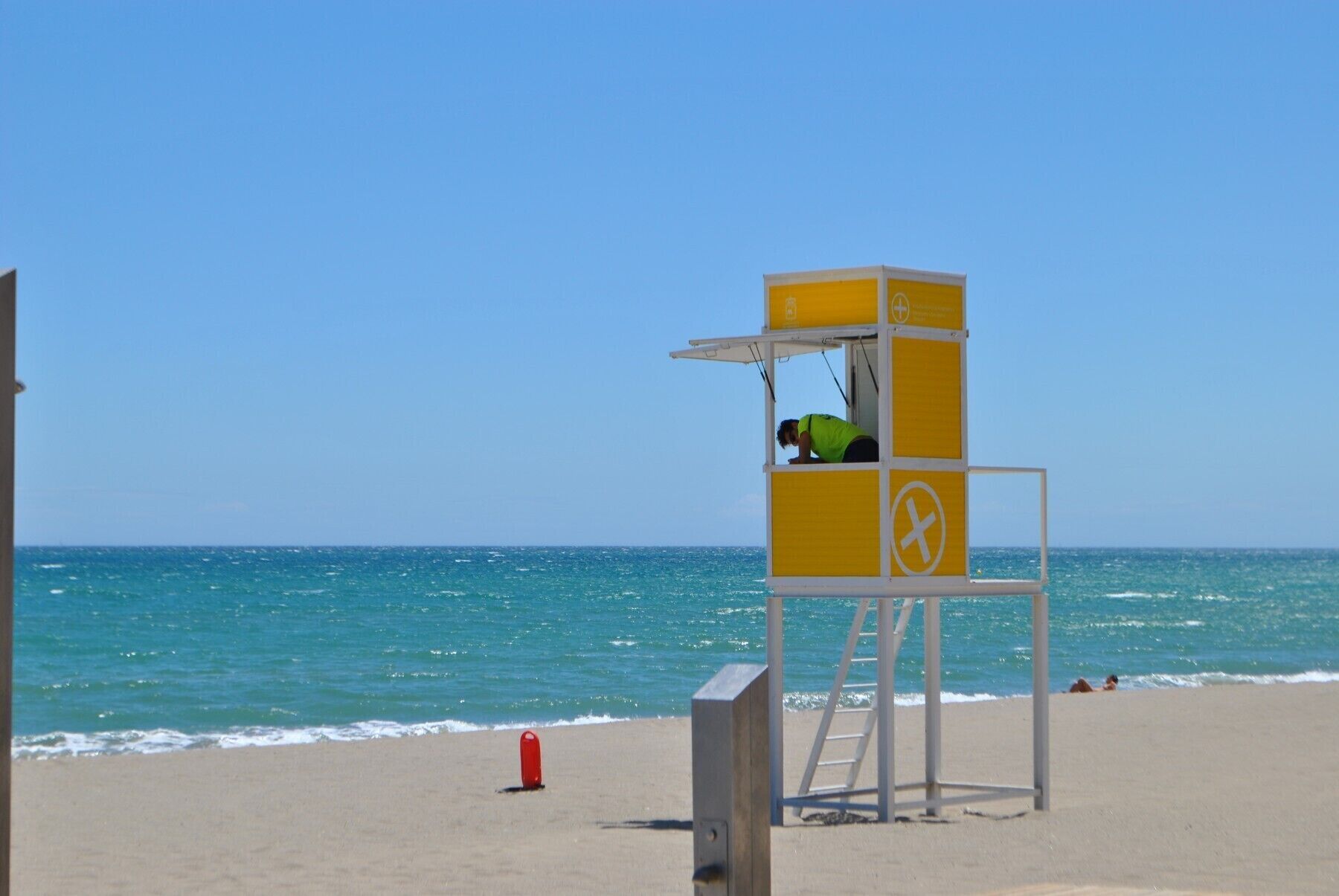 Een duikerswachthuisje met geel-witte strepen, een ladder en een rode reddingsboei aan het strand.