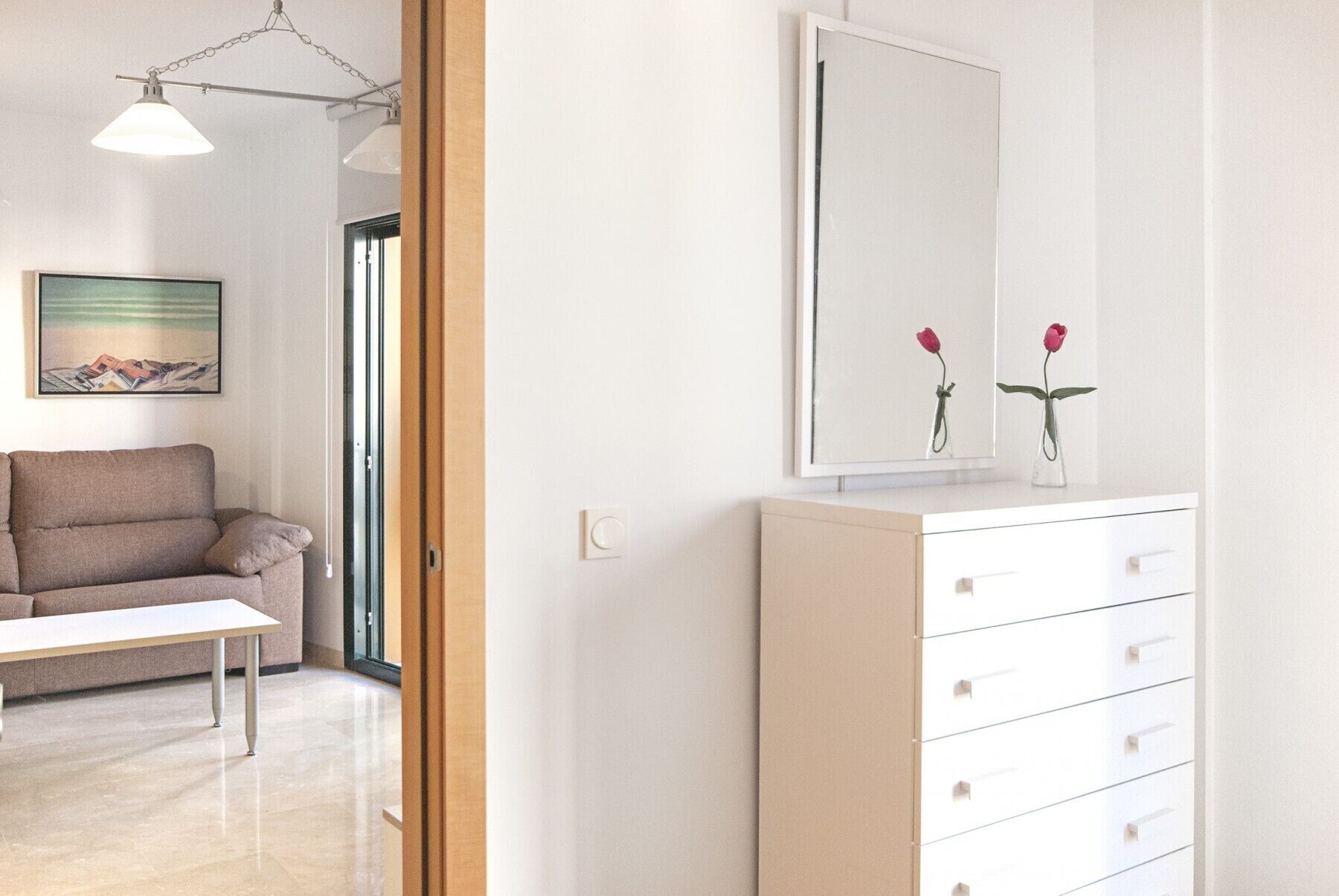 Une pièce moderne et minimaliste, avec un canapé, une commode blanche à tiroirs, un grand miroir et un tableau accroché au mur.