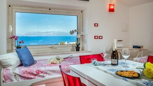 Una stanza con una grande finestra vista mare, un letto con copripiumino floreale, un tavolo da pranzo con una bottiglia di vino e un piatto di stuzzichini.