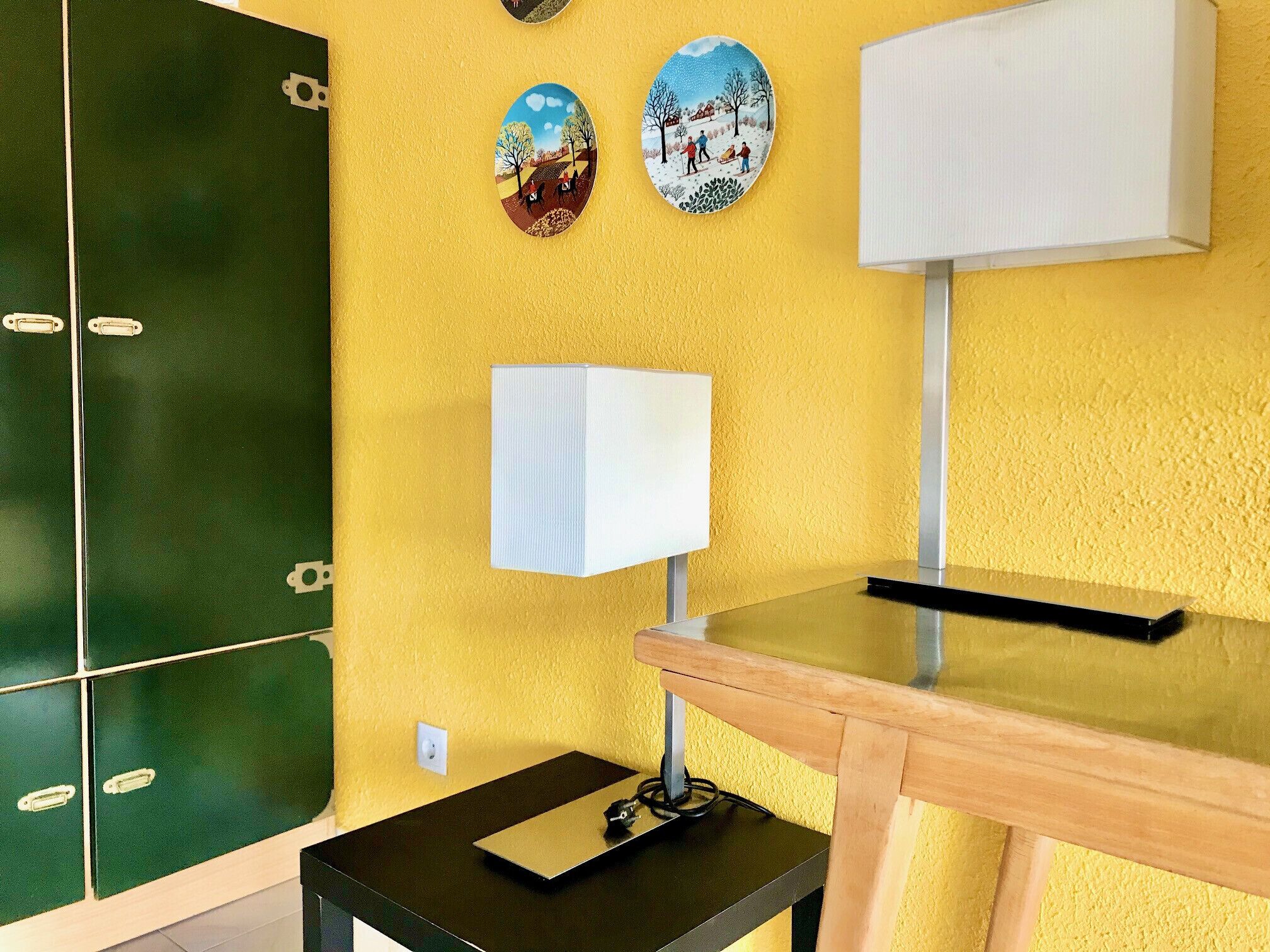 A room with a yellow wall, a green cabinet, a white lamp, and a wooden table.