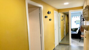 Um corredor com paredes amarelas e chão de azulejos, que leva a um quarto com cama e janela.