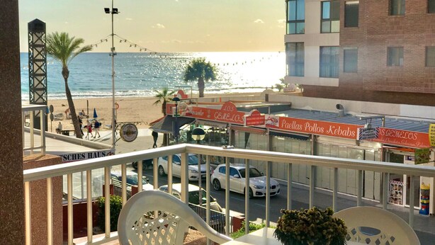 Varanda com vista para a praia, carros estacionados na rua e um anúncio que diz 'Paellas Pollo Kebabs'.
