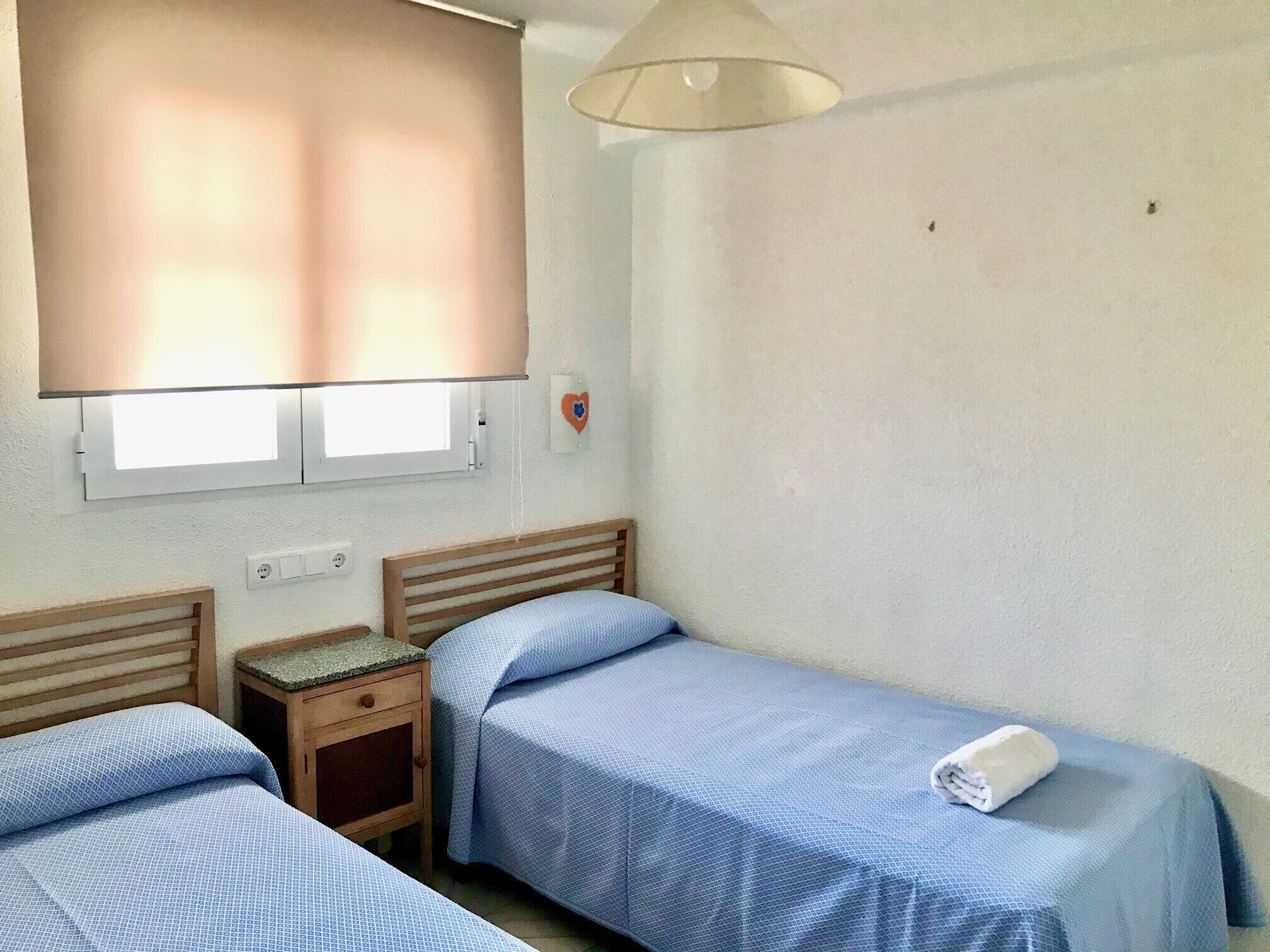Una habitación sencilla y limpia con dos camas individuales, una mesita de noche de madera, una ventana con persianas y una lámpara de pared.