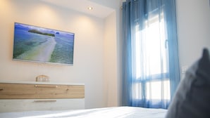 Ein Schlafzimmer mit einem Bett, einem an der Wand montierten Fernseher, einer Holzkommode und einem Fenster mit blauen Vorhängen.