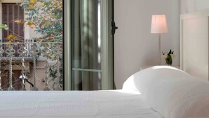 A hotel room with a bed, a nightstand, a lamp, and a view of a balcony with a railing and some foliage. - Alibei 1 - Habitat Apartments (Barcelona)