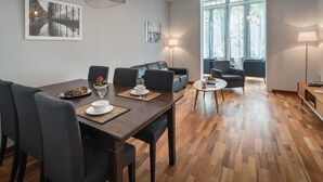 A modern living room with a dining area, featuring a wooden dining table, grey upholstered chairs, and a sofa. There are two floor lamps, a large window with curtains, and artwork on the walls. - Alibei 1 - Habitat Apartments (Barcelona)