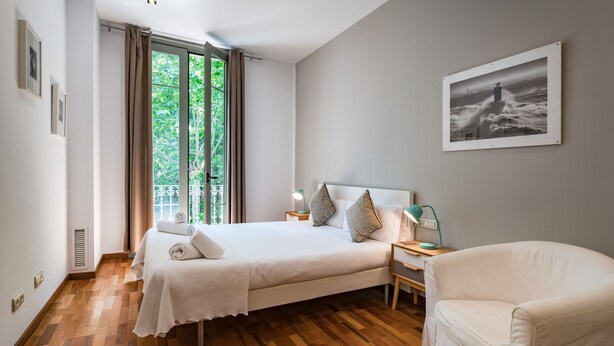 A bedroom with a bed, a nightstand, a chair, a window with curtains, and a framed picture on the wall. - Alibei 1 - Habitat Apartments (Barcelona)